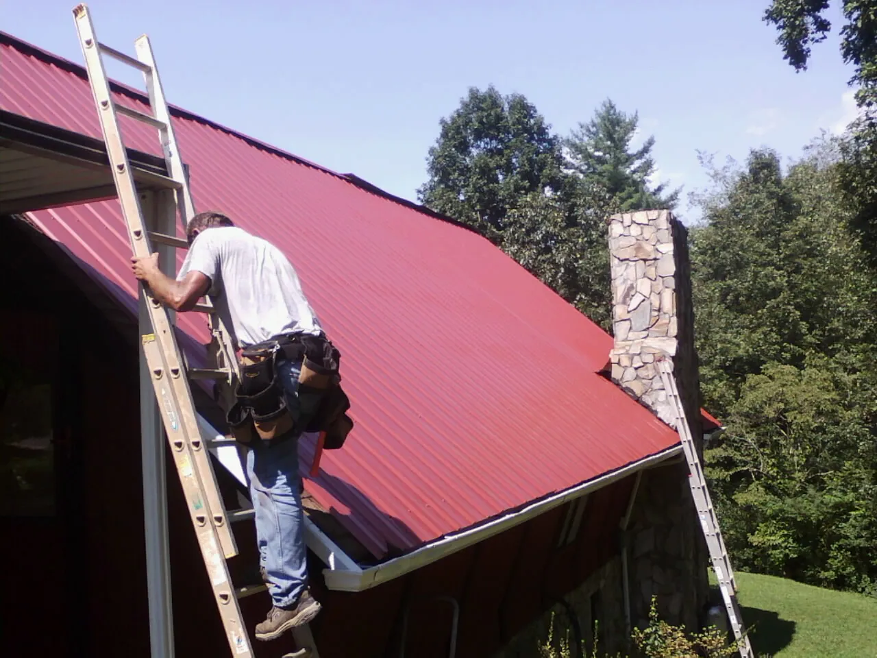 Roofer on a ladder beside a red metal roof and stone chimney — Soffit & Fascia Repair by Roofing Brothers in Ovid